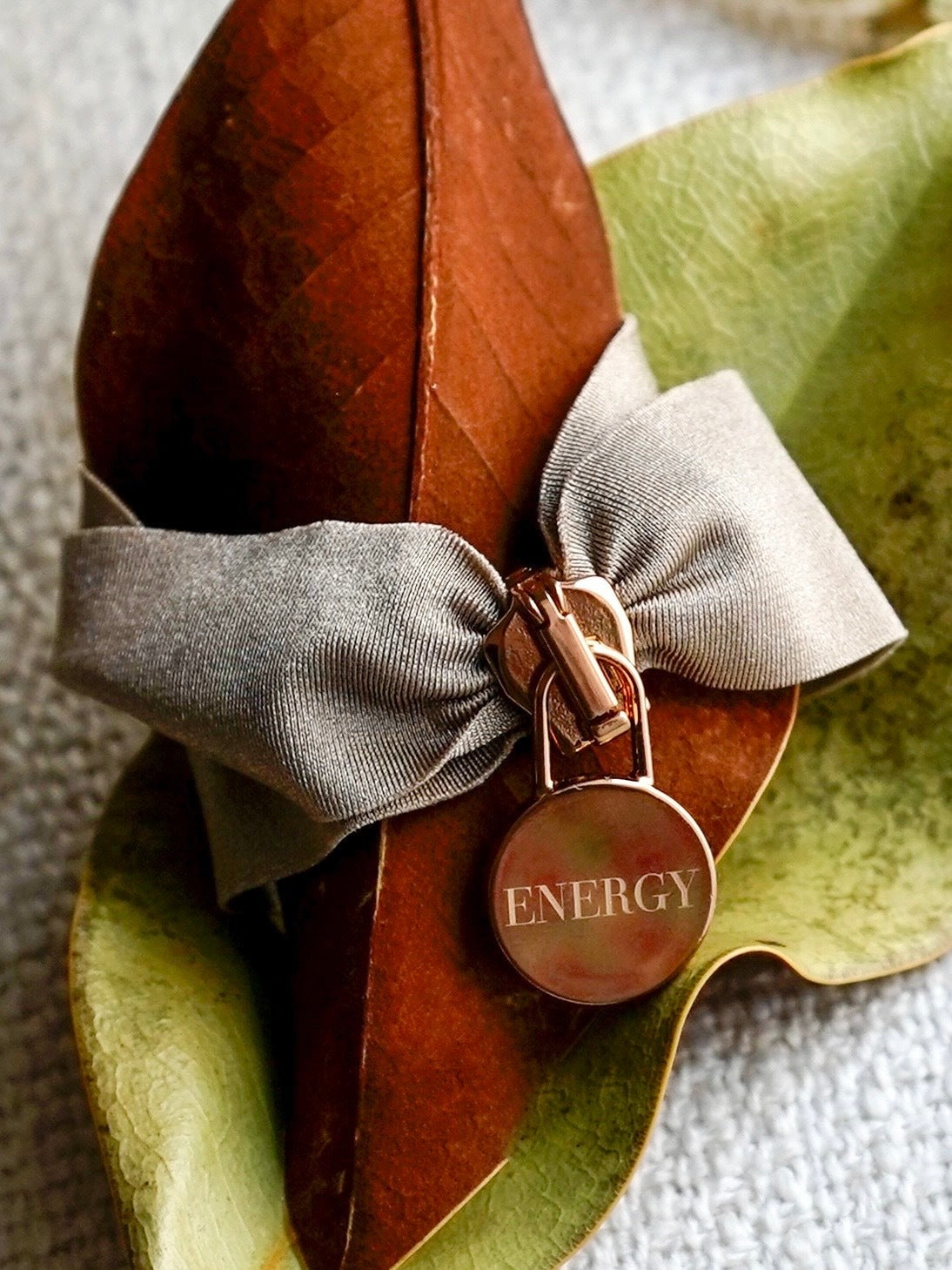 Engraved Energy Bracelet with gray bow and round ENERGY charm on natural leaves background